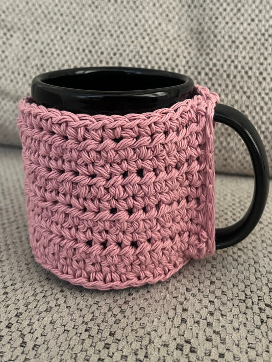 Pink crocheted mug cozy with a black handle on a textured surface